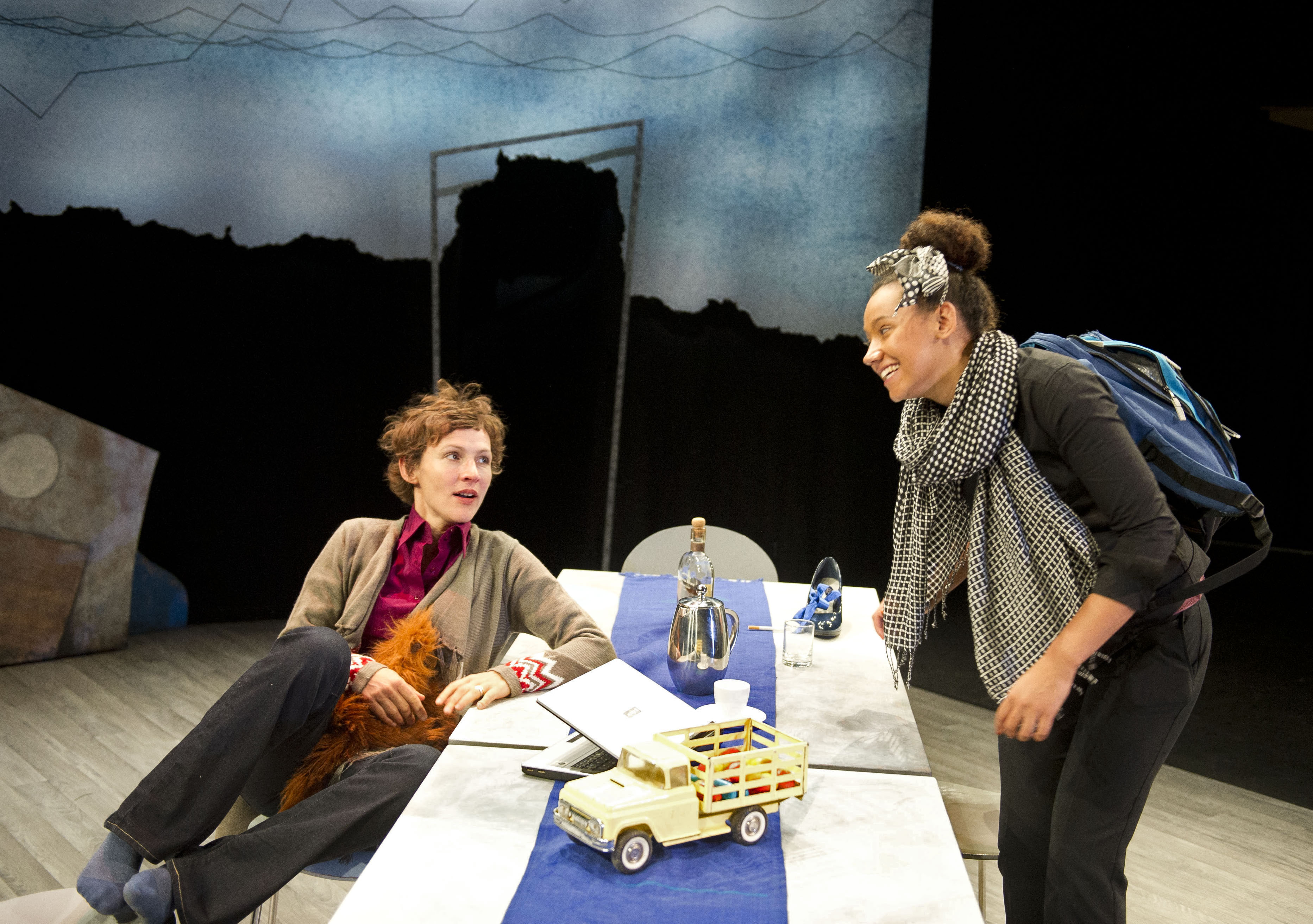 A brunette woman sits casually in a chair whilst a smiling Black woman stands beside her. On the table between them is a toy car.