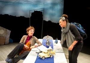 A brunette woman sits casually in a chair whilst a smiling Black woman stands beside her. On the table between them is a toy car.