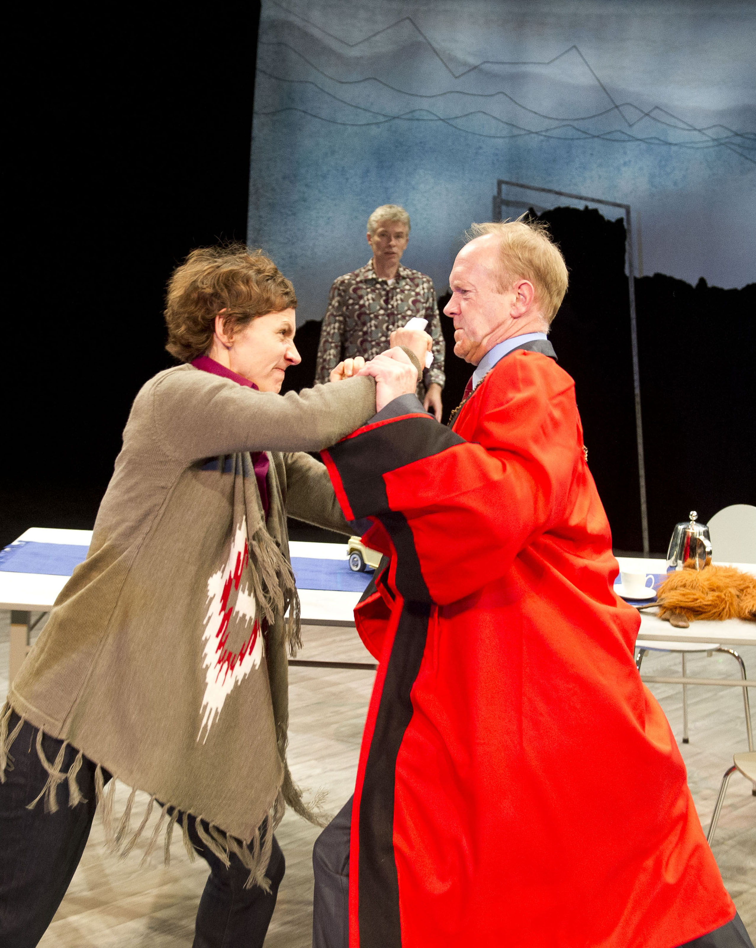 Two performers are physically fighting, faces contorted in anger. One person watches on in the background, shocked.