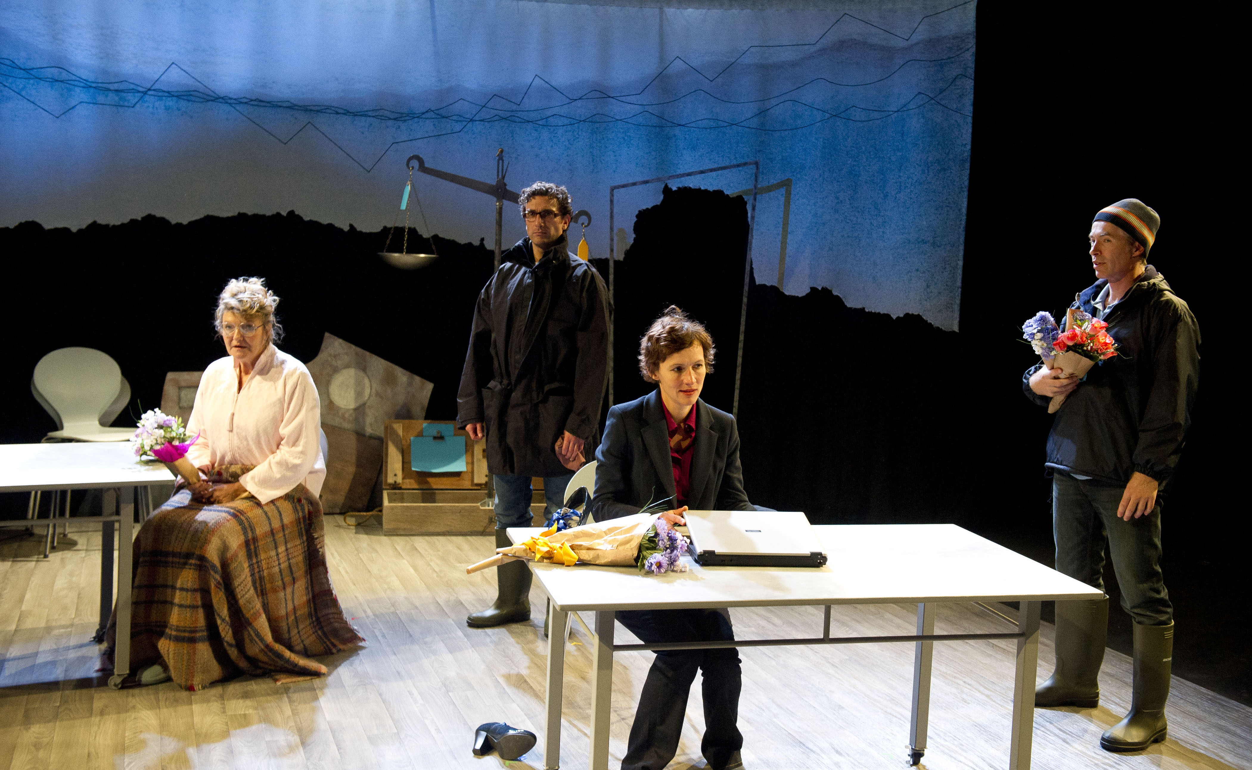 Four performers sit and stand across the stage, each absorbed in their own worlds, with a blue sky and black skyline in the backdrop behind them.