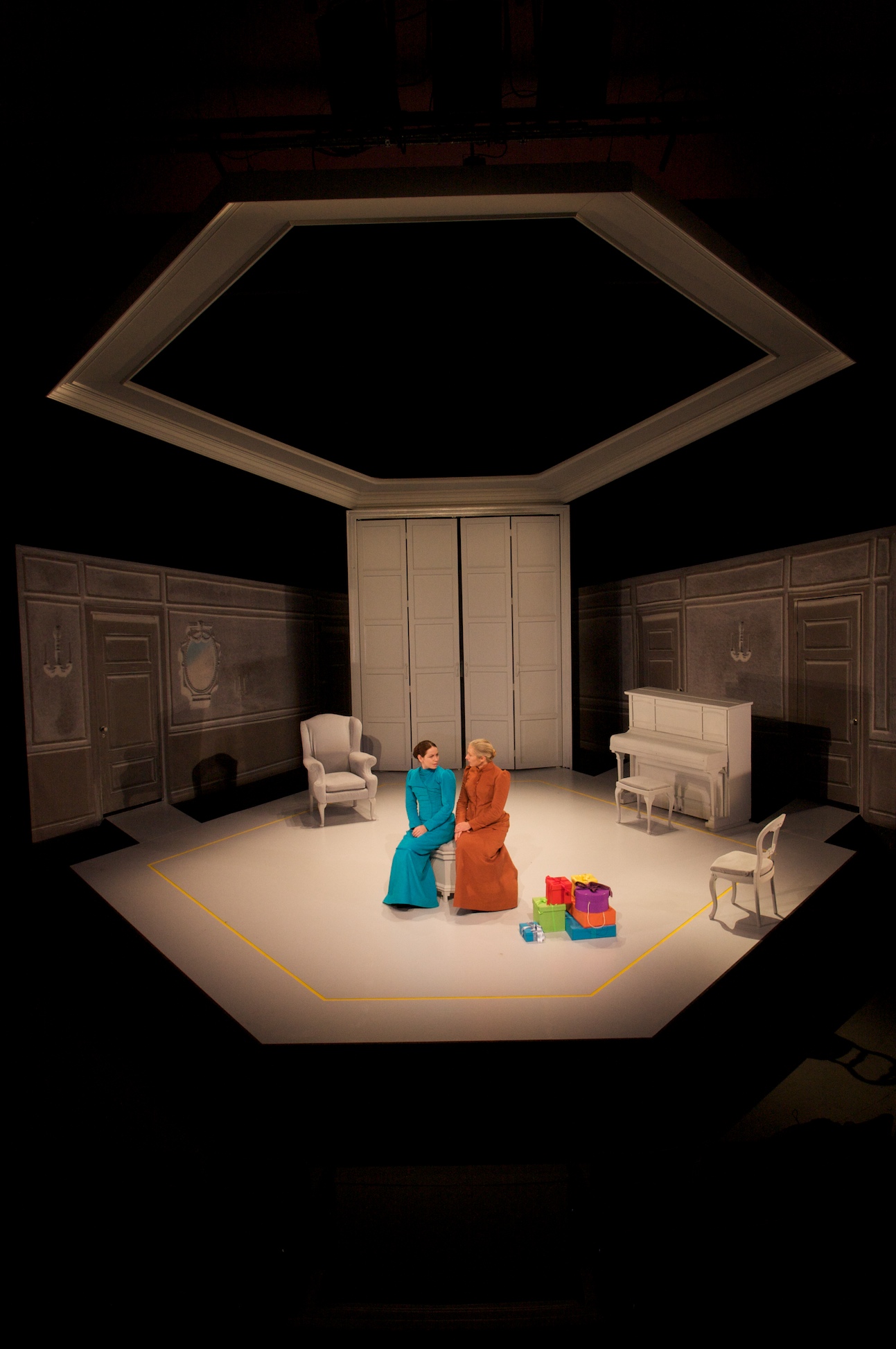 Two women sit in the middle of the stage, surrounded by a white living room and brightly coloured presents. They wear a blue and orange dress, respectively.