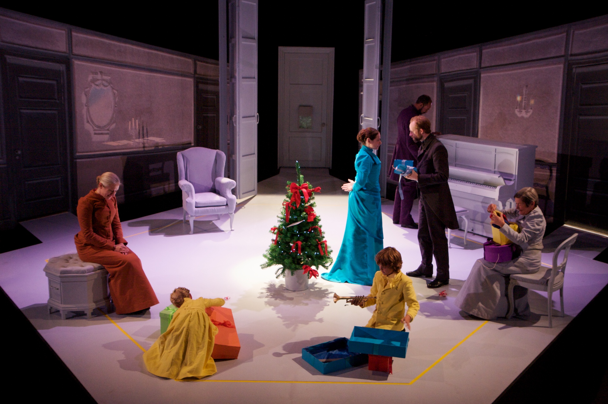 A family is seated in a period living room with a small Christmas tree in the middle. The children open presents whilst the adults talk.