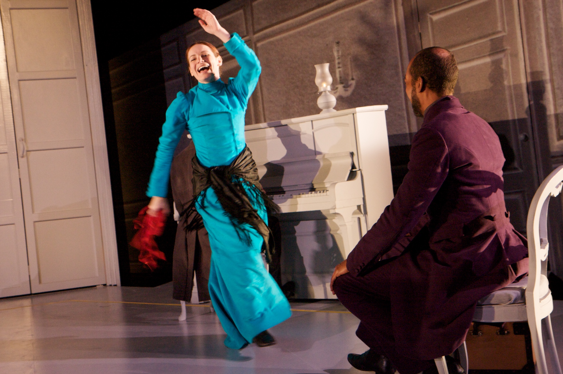 A white woman in a blue dress dances with abandon in front of a white piano. A Black man in purple period dress watches on.