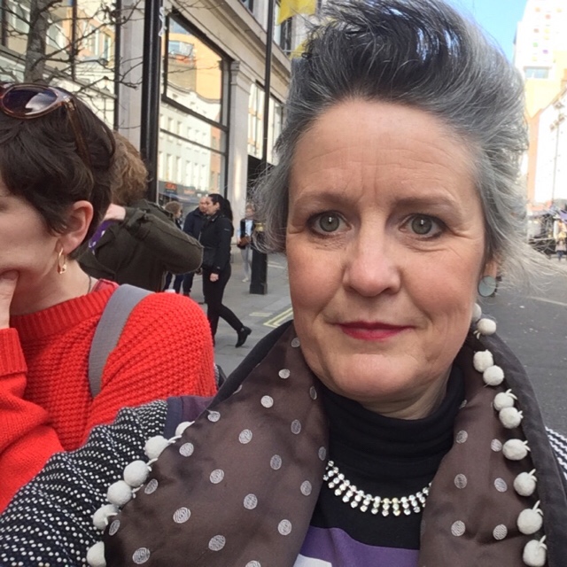 Selfie of Rebecca, a middle-aged white woman with grey and blue short hair, red lipstick and a polkadot scarf.