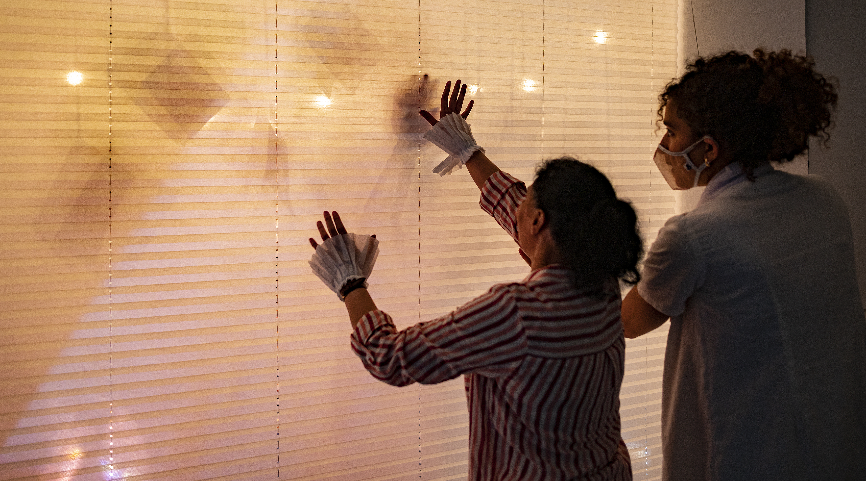 A participant reaches towards a white blind with shadows and lighting, guided by a curly-haired performer.