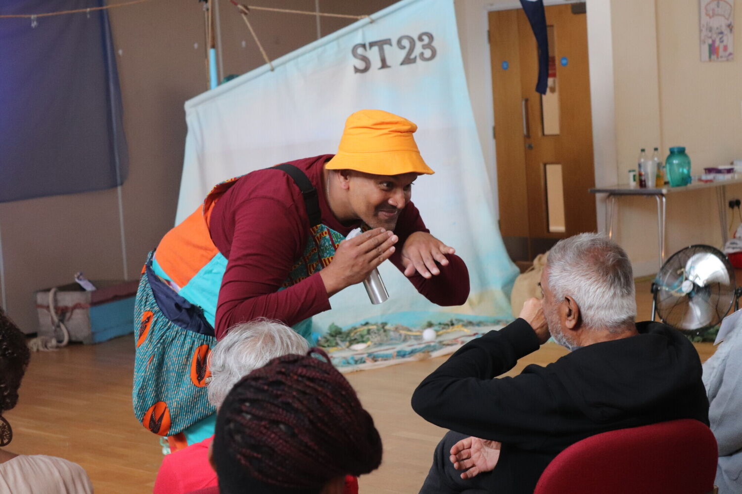 A South East Asian man in a bright yellow hat and colourful dungarees invites an audience member to smell a fragrance.