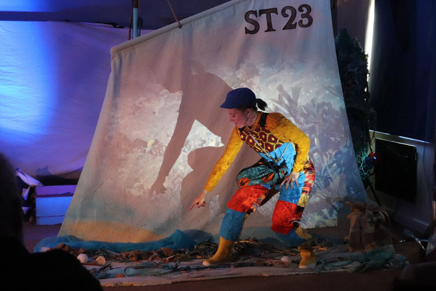 A white woman in colourful dungarees and a blue cap goes rockpooling on a theatre set, in front of a mast with a rockpool projection.